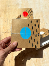 Load image into Gallery viewer, Hand holding a square cardboard product with a blue circle and black dots on a wooden surface

